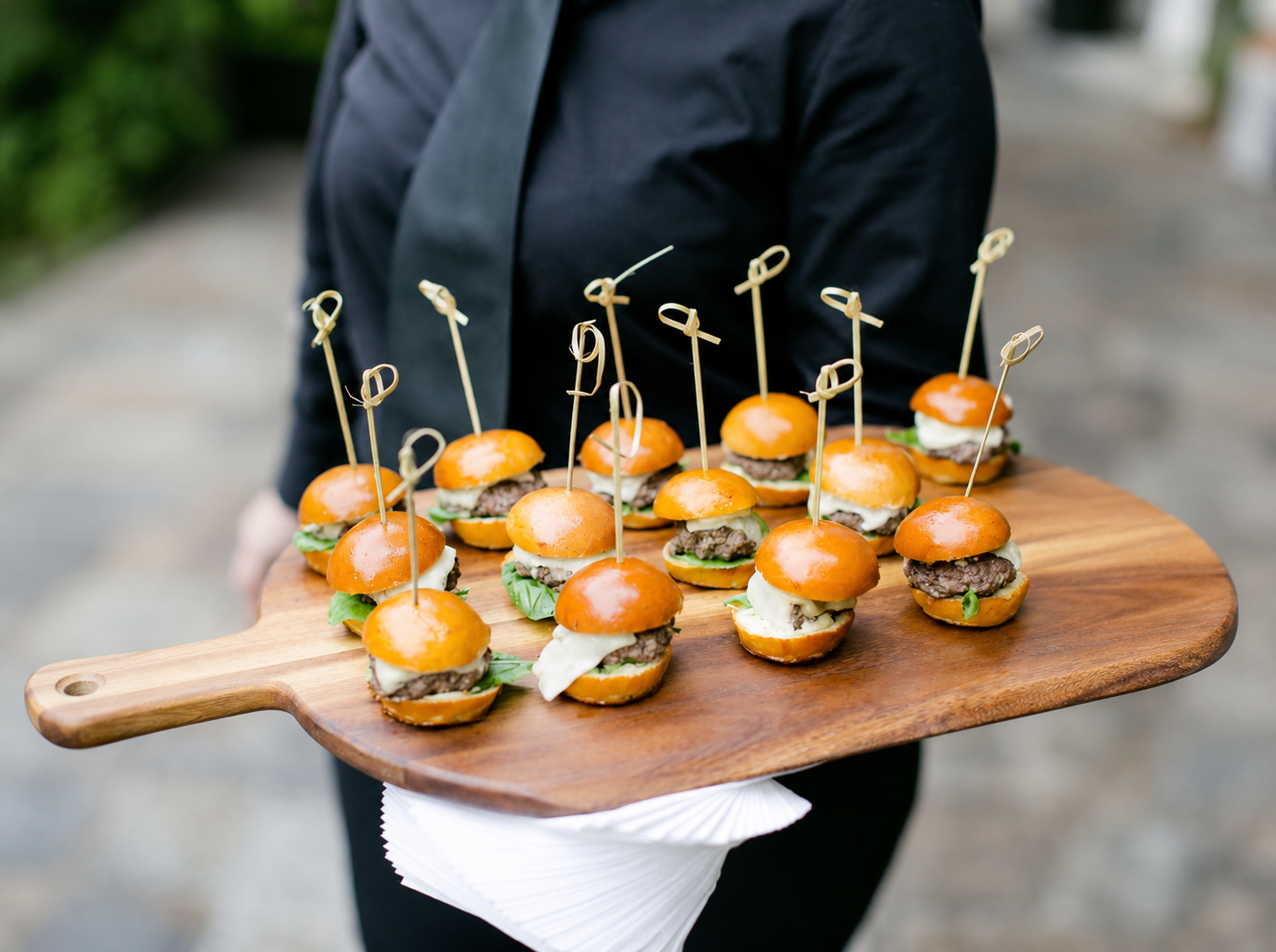 Server presenting polished mini sliders on a wooden board with bamboo picks — complete hospitality, not just delivered food